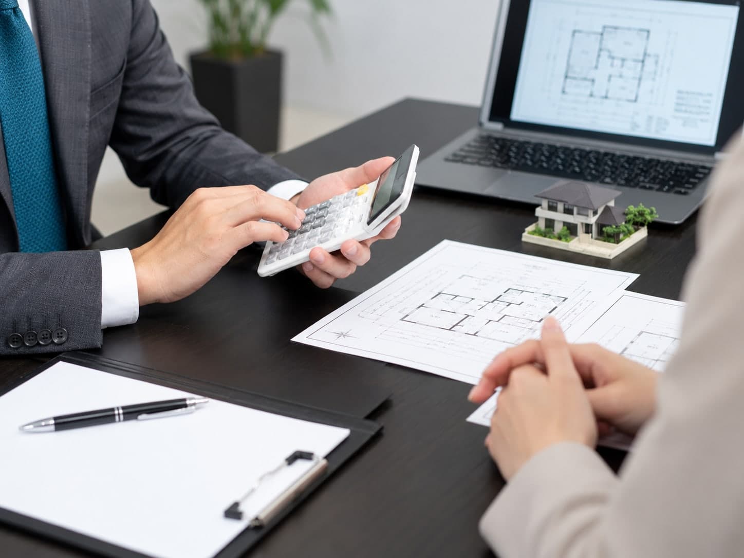 Estimator using a calculator beside floor plans, a laptop and a model house while meeting a client